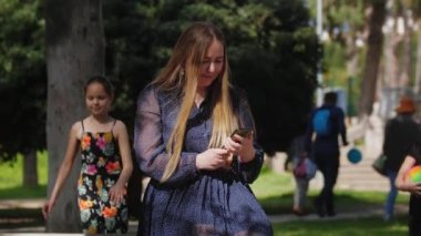 Bir kadın telefonunun ekranına bakar ve kızları ona doğru yürür. Orta çekim