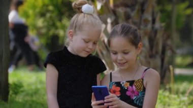 İki küçük kız kardeş çimlerin üzerinde oturup telefonla oynuyorlar. Orta çekim