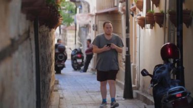 Genç bir adam sokaklarda yürüyor ve telefonuyla çevreyi vuruyor. Orta çekim