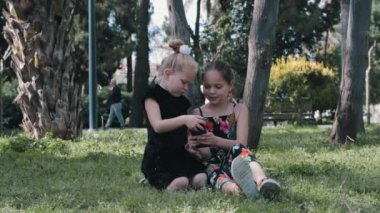İki küçük kız çimlerin üzerinde oturup telefonla oynuyorlar. Orta çekim