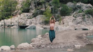 Geniş pantolonlu bir kadın gölün kenarında tek bacağıyla dengede duruyor. Orta çekim