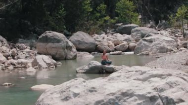 Geniş açık pantolonlu bir kadın derenin ortasındaki kayanın üzerinde meditasyon yapıyor. Orta çekim
