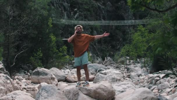 緑の森に囲まれた岩の上に立っている若い男が電話で話している ミッドショット — ストック動画