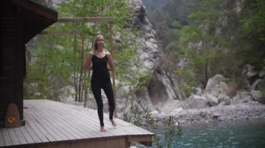 Dağlarda yoga yapan siyah elbiseli bir kadın basit jimnastik egzersizleri yapıyor. Orta çekim