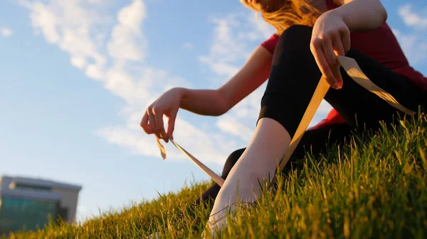 若い女性は彼女の点の靴に置き それを結びつける 日没時に緑の草の上に座っている ミッドショット — ストック写真