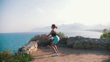 Sportif bir kadın dışarıda spor yapıyor, çömeliyor ve zıplıyor. Orta çekim