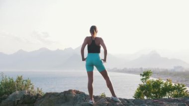 Sportif bir kadın denizin kenarındaki tepede duruyor ve ellerini uzatıyor. Orta çekim