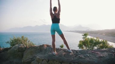 Sportif kadın deniz kenarındaki kayanın üzerinde duruyor ve ellerini kaldırıyor. Orta çekim