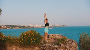 Deniz kenarındaki tepede çalışan sportif bir kadın. Olay yerinde çömeliyor. Orta çekim