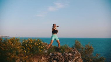 Denizin kenarındaki tepede vücudunu ısıtan sportif bir kadın. Orta çekim