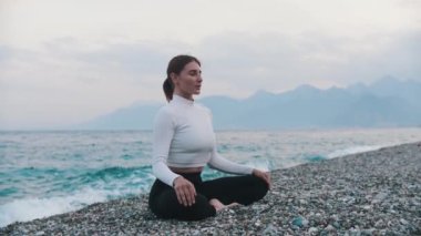 Deniz kıyısındaki çakıl taşı kumsalında nilüfer pozisyonunda oturan beyaz kazaklı bir kadın. Orta çekim