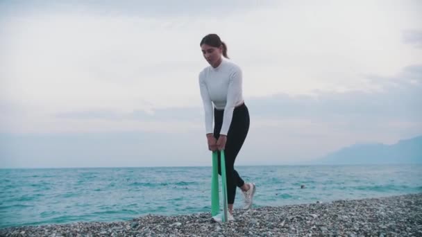 Женщина Белом Свитере Делает Упражнения Эластичной Лентой Галечном Пляже Средний — стоковое видео