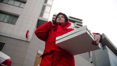 Kırmızı elbiseli bir kurye elinde pizza kutularıyla telefonda konuşuyor. Orta çekim