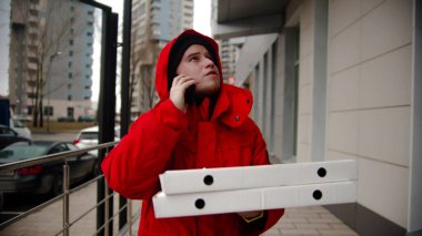 Pizza teslimatı yapan bir kurye sokakta yürüyüp telefonla konuşuyor. Orta çekim