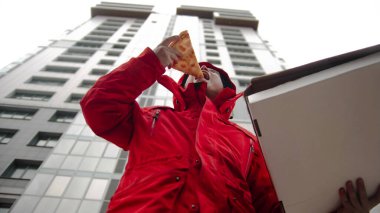 Pizza getiren bir kurye bir parça alır ve ısırır. Orta çekim