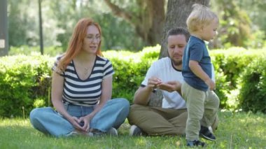Aile parkta vakit geçirir. Aileler oğullarının çimenlerin üzerinde oturur ve onlara koparılmış çimen verir. Orta çekim