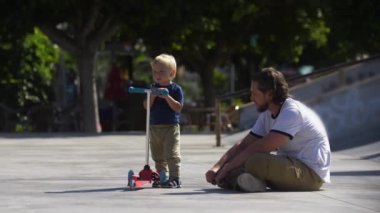 Father with his little son in the skatepark about to traing skating on the scooter. Mid shot