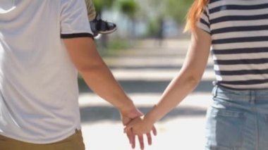 Young parents holding hands and walking in the green park. Mid shot