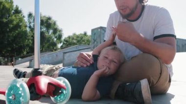 Little boy with his father sitting on the floor in the skate park. Mid shot