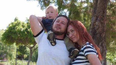 Aile, yeşil bir parkta vakit geçirir. Küçük sarışın çocuk babasının omuzlarında oturur. Orta çekim
