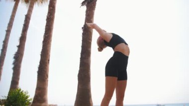 Deniz kenarındaki jimnastik salonunda plastik sarışın bir kadın ayağa kalkar ve köprü pozuna doğru eğilir. Orta çekim