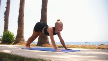 Deniz kenarında jimnastik yapıyor. Plastik sarışın kadın sırtı için egzersiz yapıyor ve yoga minderinde omurga çalışıyor. Orta çekim