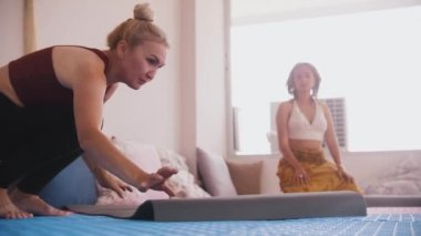Older and younger women in the yoga studio - spreads out a yoga mat and gets into a pose. Mid shot