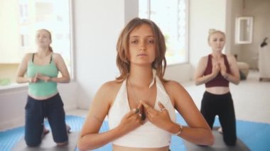 Three women having yoga classes in the studio - the younger one stands in the front and running her hands above her chest. Mid shot