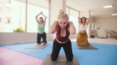 Three women having yoga classes in the bright studio - the older one stands in the front and all of them bending down to the floor. Mid shot