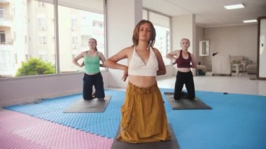 Three women having yoga classes in the studio - the younger one stands in the front and all of them running their hands against their bodies. Mid shot