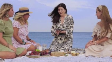 Four adult women at a picnic by the sea playing a card game. Mid shot