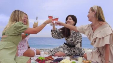 Four women clink glasses of wine at a picnic by the sea. Mid shot