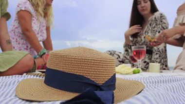 Women having picnic on the beach - one of them picks up a straw hat. Mid shot