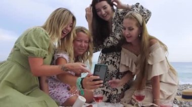 Four excited women watching funny videos on the phone while having picnic by the sea. Mid shot