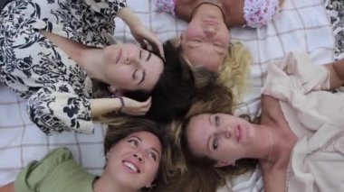 Four women lie with their heads on a blanket and talk to each other. Mid shot
