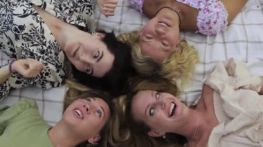 Four women lie with their heads on a blanket and talking. Mid shot