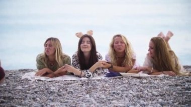 Beach picnic - four women lying on the blanket and talking by the sea. Mid shot