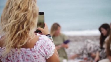 A woman takes pictures on the phone of her friends on the beach by the sea. Mid shot