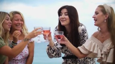 Four smiling women clink glasses of alcohol on the beach. Mid shot