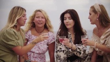 Four women friends drinking wine on the beach by the sea. Mid shot