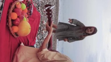 A woman playing guitar by the sea and her little daughter dancing and picks up a berry from a plate on the blanket. Mid shot