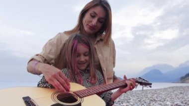 Beautiful woman teaching her little daughter playing guitar on nature. Mid shot