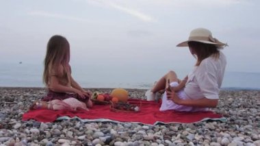 Woman filming her little daughter on her phone while having a picnic by the sea. Mid shot