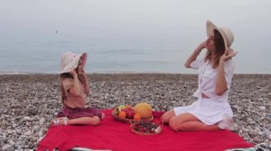 Family of mother and her little daughter having a picnic by the sea and trying on each others hats. Mid shot