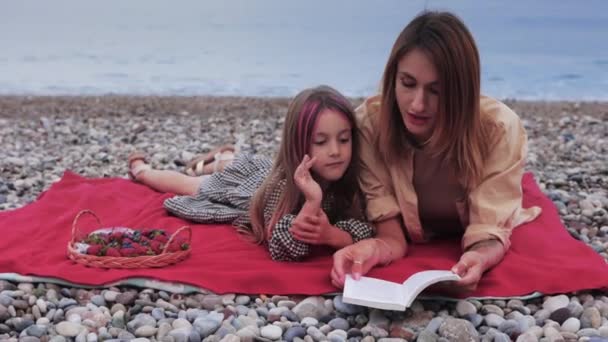 海でピクニックをしながら本を読んでいる母親を持つ少女 ミッドショット — ストック動画