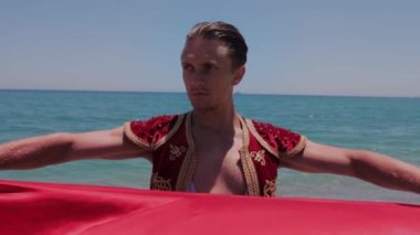 Çekici bir dansçı deniz suyunda kırmızı bir manto tutuyor ve yana bakıyor. Orta çekim