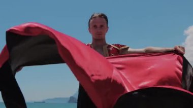 Ulusal takım elbiseli çekici bir dansçı kumsalda duruyor ve elinde kırmızı manto var. Orta çekim