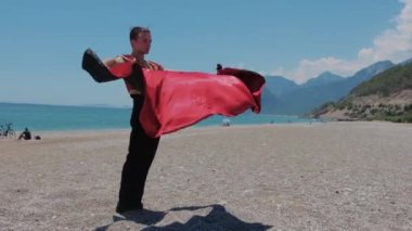 Sahilde duran ve elinde kırmızı manto tutan sahne kostümlü çekici bir adam. Orta çekim
