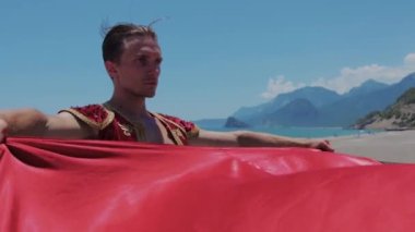 Sahilde kırmızı manto tutan sahne kostümlü çekici bir adam. Orta çekim