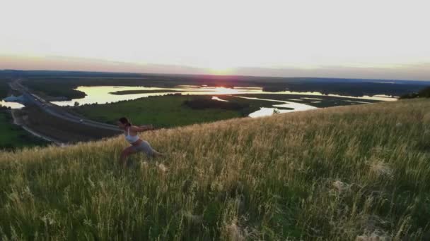 日没時に高速道路の上の緑の丘の上でスポーツ演習を行う一人の女性 空中風景 — ストック動画